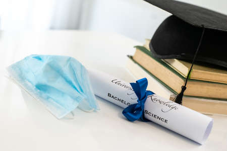 Closeup of graduation hat with degree and surgical mask on white background. On line virtual celebration at home for students during pandemic coronavirus. Graduation in lockdown. Graduating concept.の写真素材