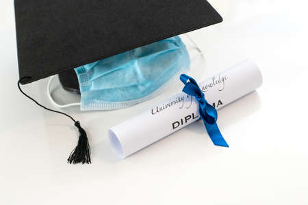 Closeup of graduation hat with degree and surgical mask on white background. On line virtual celebration at home for students during pandemic coronavirus. Graduation in lockdown. Graduating concept.の写真素材