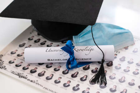 Closeup of graduation hat with degree and surgical mask on white background. On line virtual celebration at home for students during pandemic coronavirus. Graduation in lockdown. Graduating concept.の写真素材
