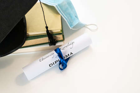 Closeup of graduation hat with degree and surgical mask on white background. On line virtual celebration at home for students during pandemic virus. Graduation in lockdown. Graduating concept.の写真素材