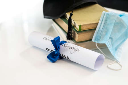 Closeup of graduation hat with degree and surgical mask on white background. On line virtual celebration at home for students during pandemic virus. Graduation in lockdown. Graduating concept.の写真素材