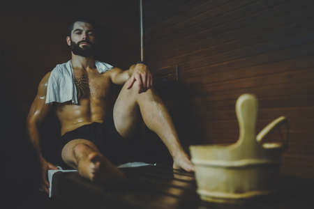 Beautiful healthy boy relaxing in a woden sauna. Young bearded man resting after workout in the gym....