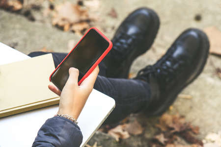 Girl with wool cap sitting in the park using smartphone, beautiful autumn afternoon. Teen using mobile phone, chat with friends and classmates. Young woman enjoying free time in nature with laptoの写真素材