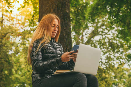 Girl study sitting on a bench in the park in a beautiful autumn afternoon. Teen using laptop, smartphone and reading a book outdoor. Student alone doing homework outside in the nature. School conceptの写真素材