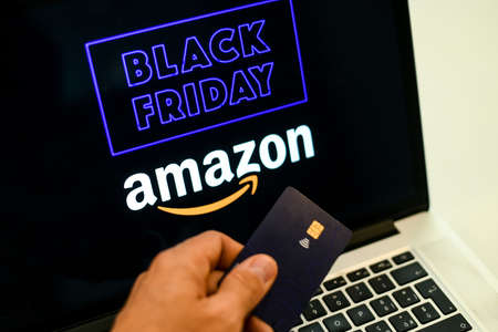 Hand with credit card in front of the laptop monitor doing purchases Amazon on line shop black friday. Purchase Christmas gifts. E-commerce, tech, online shopping concept Milan, Italy - November 2021のeditorial素材
