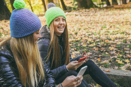 Two girls with wool cap sitting in the park using smartphone, beautiful autumn afternoon. Teen using mobile phone. Sisters chat with friends and classmates. Young women enjoying free time in natureの写真素材