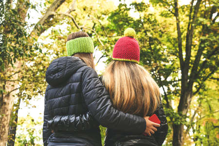 View from the back of two girls in the park. Teen walking outdoor in the forest. Young student woman enjoying free time in nature. Women embraced in a beautiful autumn afternoon looks the trees.の写真素材