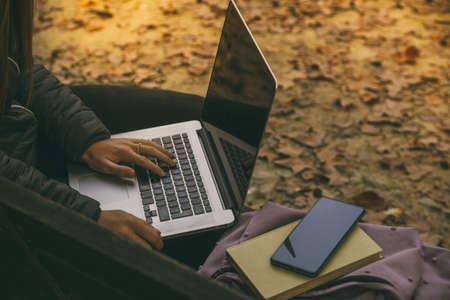 Girl study sitting on a bench in the park in a beautiful autumn afternoon. Teen using laptop, smartphone and reading a book outdoor. Student alone doing homework outside in the nature. School conceptの写真素材