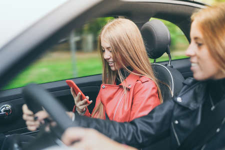 Trendy girls sitting in the car. Smiling teen in front of the steering wheel with a friend using...