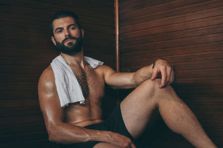 Beautiful healthy boy relaxing in a wooden sauna. Young bearded man resting after workout in the gym. Fitness male with tattoo and white towel, laying on the bench in the spa. Wellness, health conceptの写真素材