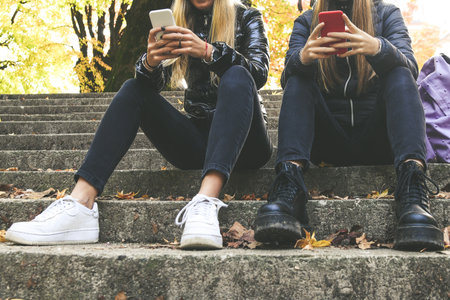 Two girls with wool cap sitting in the park using smartphone, beautiful autumn afternoon. Teen using mobile phone. Sisters chat with friends and classmates. Young women enjoying free time in natureの写真素材