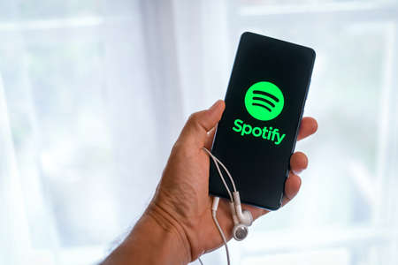 Close up view of hand with smartphone and Spotify logo on display. Window on background. New...