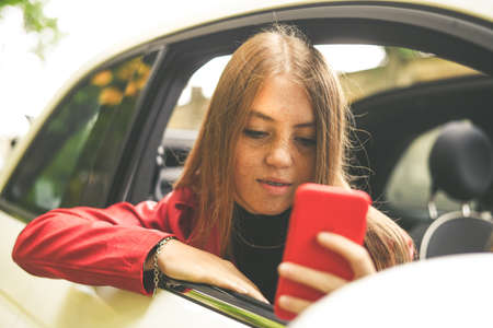 Trendy girl sitting in a convertible car with security belt. Smiling teen using smartphone and chat...