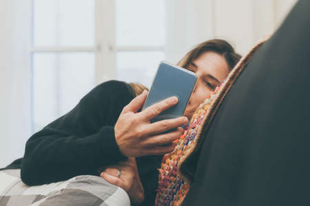 Relaxed girl using smartphone sitting on the sofa at home. Woman chatting with friends sitting on a couch in the living room with warm pajama in a winter morning. Leisure, carefree and relax concept.の写真素材