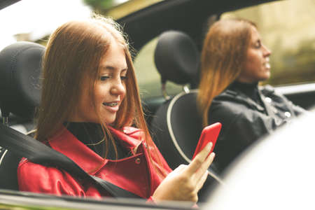 Trendy girls sitting in the car. Smiling teen in front of the steering wheel with a friend using...