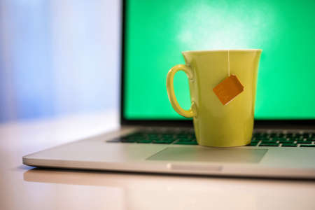 Closeup view of a cup of hot beverage and a laptop with green screen in background. Steaming yellow mug on computer keyboard. Remote workstation. Smart working, home working and homeschooling conceptの写真素材