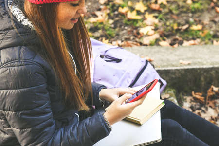 Girl with wool cap sitting in the park using smartphone, beautiful autumn afternoon. Teen using mobile phone, chat with friends and classmates. Young woman enjoying free time in nature with laptoの写真素材
