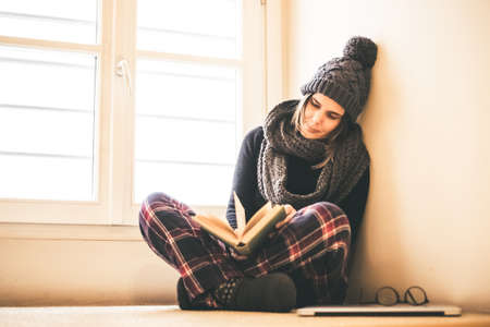 Young beautiful girl reading a book during cold winter with wool cap and scarf to keep warm. Woman...