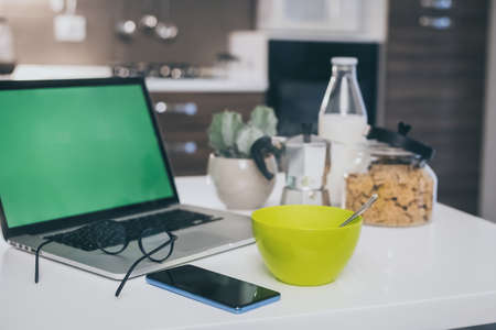 Table set for breakfast, kitchen in the background. Natural meal and technology devices. Interior view of a cozy and modern home. Millennial, youth, home working, home schooling, remote work concept.の写真素材