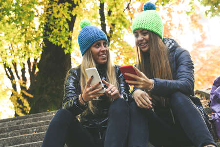 Two girls with wool cap sitting in the park using smartphone, beautiful autumn afternoon. Teen using...