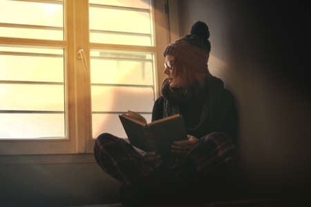 Young beautiful girl reading a book during cold winter with wool cap and scarf to keep warm. Woman sit read near the window. Female relaxing at home. Lifestyle lockdown carefree and education conceptの写真素材