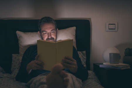 Man alone in pajamas reading a book lying on bed at late at night. Boy sitting in bedroom reads a novel at home. Male enjoy free time at home in relaxation. Lifestyle, bedtime, rest and people conceptの写真素材