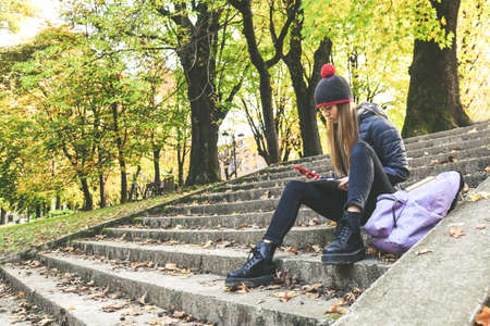 Girl with wool cap sitting in the park using smartphone, beautiful autumn afternoon. Teen using...