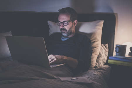 Man works with laptop lying on bed. Male home working with remote office. Boy sitting in bedroom use online devices. Smart working, home school, internet lifestyle new communication technology conceptの写真素材