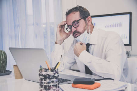 Fatigued business man with surgical masks working during lockdown for coronavirus covid-19. Exhausted male sitting at the desk works with laptop. Health care, quarantine, remote working concept.の写真素材