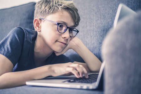 Young student doing homework lying on a sofa using laptop. Caucasian boy watching video online alone at home. Teen enjoying free time with computer. New technology education concept Soft focus on eyesの写真素材