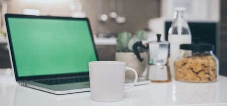 Table set for breakfast, kitchen in the background. Natural meal and technology devices. Interior view of a cozy and modern home. Millennial, youth, home working, home schooling, remote work concept.の写真素材