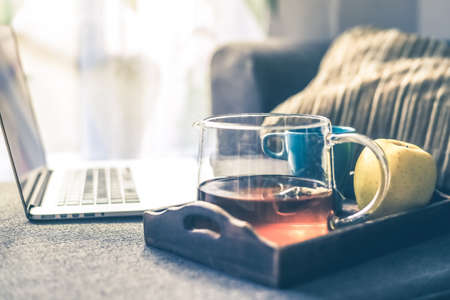 Still life of a natural breakfast at home beside a laptop on the sofa. Carafe of tea, coffee and an apple on a wooden tray with a computer. Home work, new trend technology and healthy food concept.の写真素材