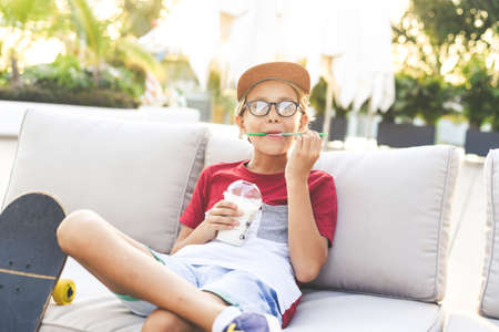 Trendy young boy drinking a smoothie sitting outdoor at the bar. Teen with hat drinks a cold...