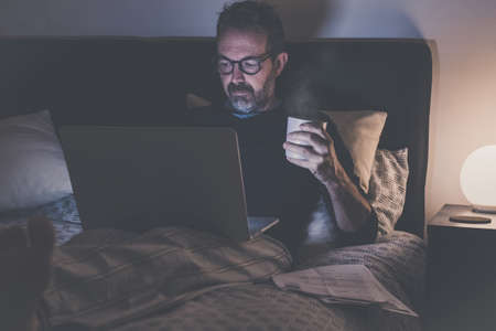 Man works with laptop lying on bed. Male home working with remote office. Boy sitting in bedroom use...