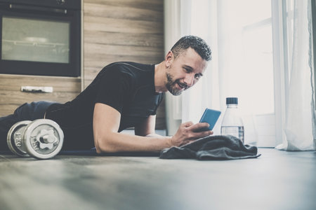Sporty man resting after working out at home. Fit boy looking gym exercise video on smartphone....
