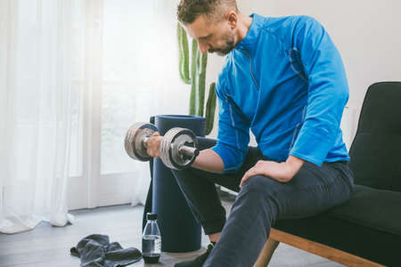 Determined sporty young man working out and trains at home. Fit male doing exercise with weights for biceps. Strong boy lifting the dumbbells in the living room. Wellness, health, sport, gym conceptの写真素材