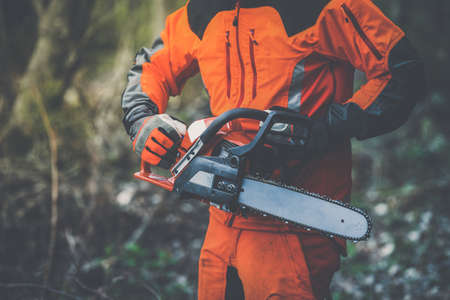Close up view of an hand holding a chainsaw. Lumberjack at work wears orange personal protective equipment. Gardener working outdoor in the forest. Security, professionalism, occupation worker conceptの写真素材