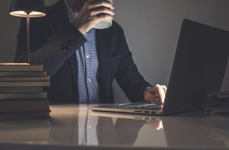 Businessman working with laptop mug in hand. Documents, books on the desk. Warm, intimate atmosphereの写真素材