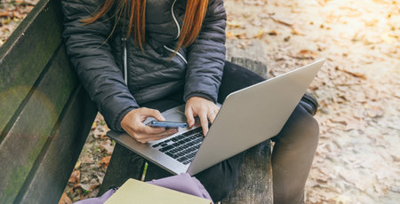 Girl study sitting on a bench in the park in a beautiful autumn afternoon. Teen using laptop, smartphone and reading a book outdoor. Student alone doing homework outside in the nature. School conceptの写真素材
