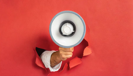 Hand holds a white megaphone breaks through a red wall. Concept of communication, message, announceの素材