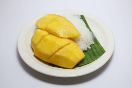 Most popular Thai  dessert, ripe yellow mango with sticky rice served with coconut milk  on banana leaf on white plateの写真素材