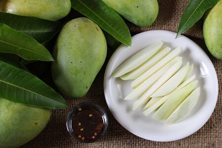 Sliced tropical fresh green mango serve with sweet fish sauce, top view with green mangoes and green leaves on burlap sackの写真素材