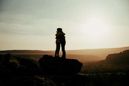 Silhouette caucasian husband and wive hugging on top of mountain at sunset while on honeymoonの写真素材