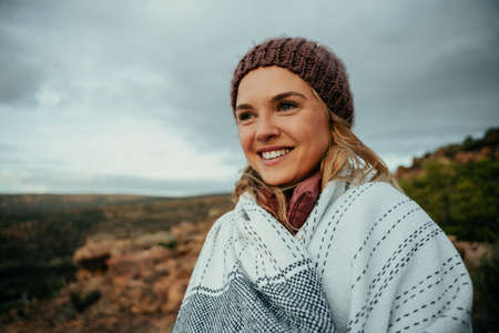 Caucasian female wearing beanie hugging warm blanket smiling while standing on top of mountain