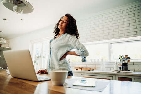 Mixed race female student tired standing to stretch sitting in kithcen at laptopの写真素材