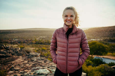 Caucasian blonde female smiling with hands in pocket standing on mountain topの写真素材