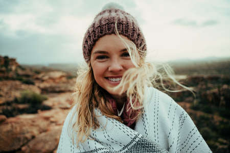 Caucasian female smiling wearing beanie wrapped in cozy blanket enjoying camping on mountain topの写真素材