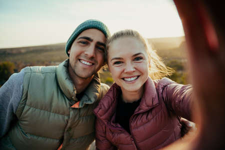 Caucasian husband and wife taking selfie standing on top of mountain smiling while sun sets behindの写真素材