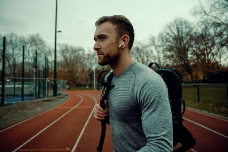 Caucasian male athlete walking on track field holding backpack wearing earphones