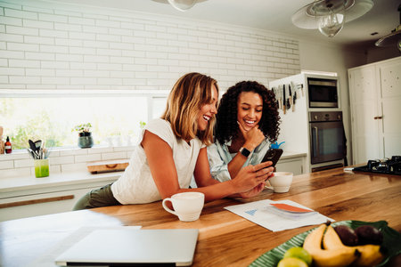 Mixed race female students taking work break chatting in kitchen with cellular device and hot coffeeの写真素材